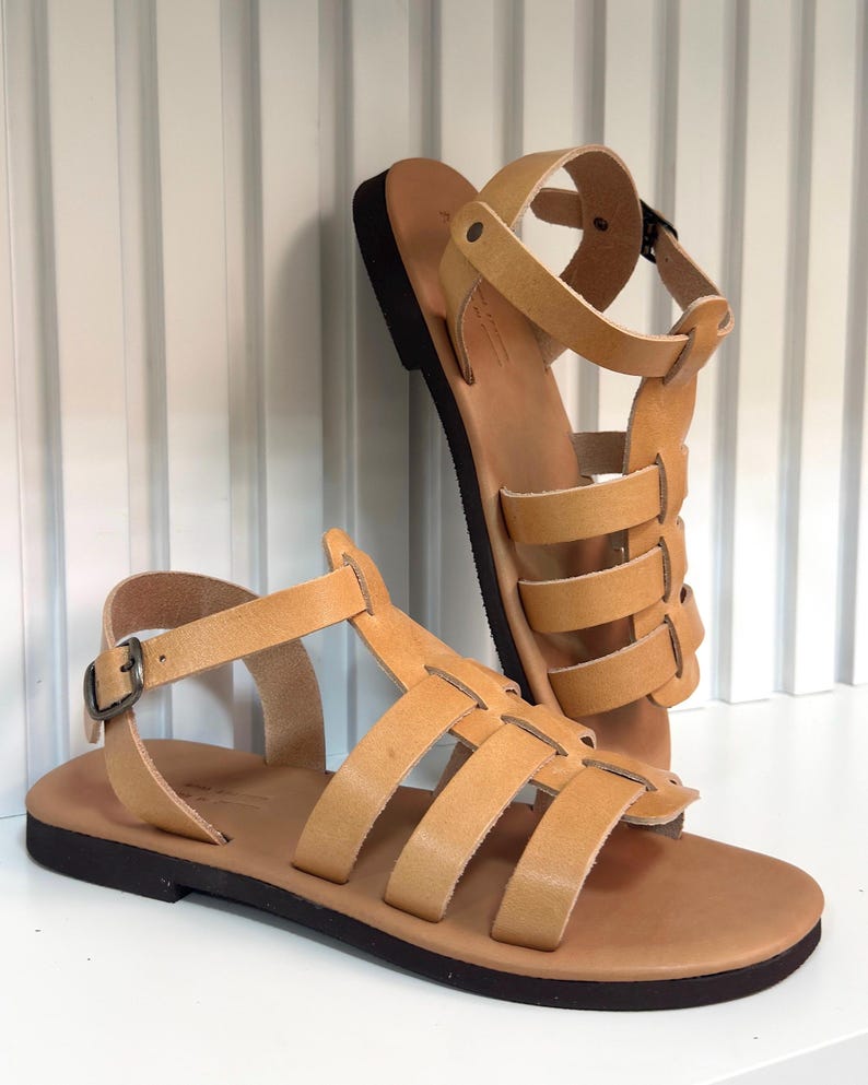 Pu&ograve; includere: Un paio di sandali in pelle color cuoio con pi&ugrave; cinturini e chiusura con fibbia. I sandali hanno una suola marrone scuro e sono esposti su una superficie bianca. Il design ricorda le calzature romane antiche.