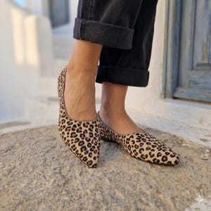 Peut inclure: Une paire de chaussures plates à bout pointu avec un motif imprimé léopard beige et noir. Les chaussures présentent une silhouette décolletée et un profil fin, portées avec un pantalon noir à revers s'arrêtant juste au-dessus de la cheville.