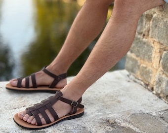 Puede incluir: Sandalias de cuero marrón con diseño de tiras. Las sandalias tienen una suela marrón claro y cierre de hebilla. La imagen muestra las sandalias en los pies de una persona, sugiriendo un tema casual y al aire libre.