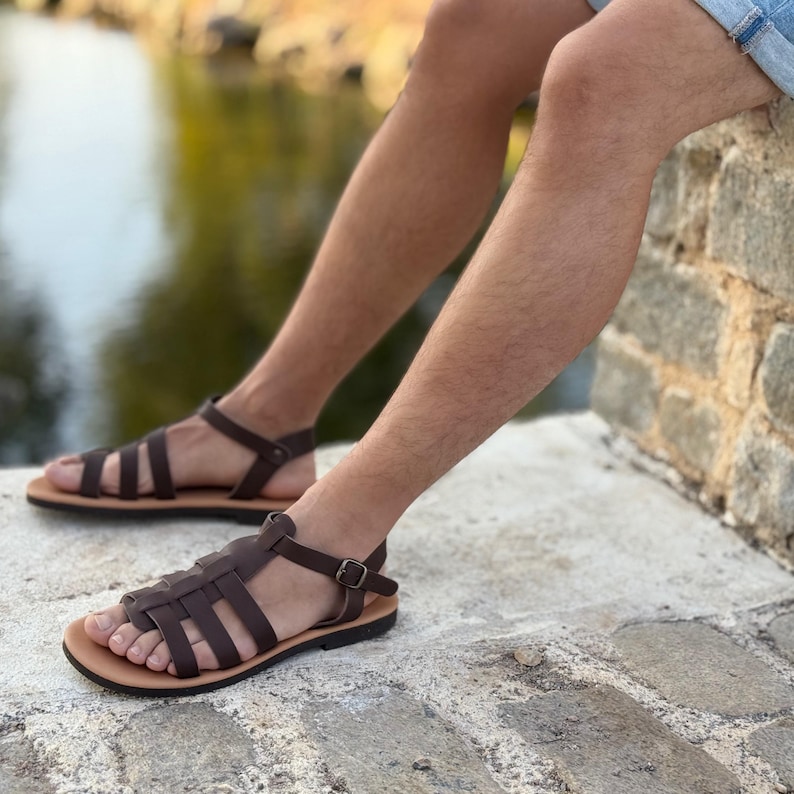 Brown leather sandals with a strappy design. The sandals have a light brown sole and a buckle closure. The image shows the sandals on a person's feet, suggesting a casual, outdoor theme.