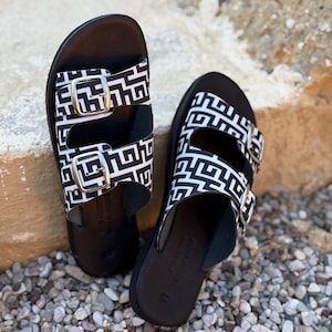 Puede incluir: Un par de sandalias negras con un patrón geométrico blanco y negro en las correas. Las sandalias tienen dos correas ajustables con hebillas plateadas. Las suelas son negras y las sandalias están sobre pequeñas piedras grises y marrones.