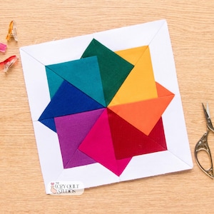 Könnte beinhalten: Ein quadratischer Quiltblock mit einem leuchtenden Sternmuster aus türkisfarbenen, dunkelgrünen, gelben, orangen, roten, magentafarbenen, violetten und blauen Stoffdreiecken, umrahmt von einem weißen Rand, mit drei kleinen Clips und einer Stickschere.