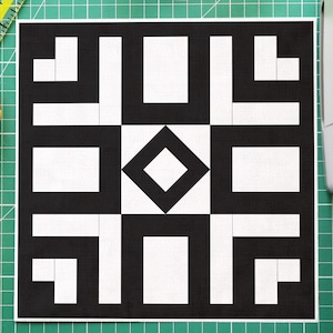 May include: A black and white geometric quilt block design on a green cutting mat. A yellow rotary cutter, a clear bowl of colourful pins, a clear ruler, and a sewing machine are also visible.