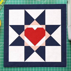 May include: A handmade quilt block with a red heart center, surrounded by white, navy, and pink geometric shapes. A yellow rotary cutter, a green cutting mat, a clear bowl of pins, and a ruler are also visible.