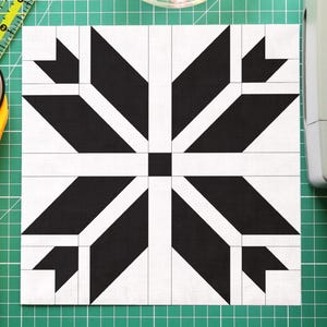 Puede incluir: Un bloque de edredón geométrico en blanco y negro con un diseño de copo de nieve está centrado en una esterilla de corte verde. Un cortador rotatorio amarillo y un cuenco transparente con alfileres de colores están junto al bloque. Una regla transparente está en la parte superior.