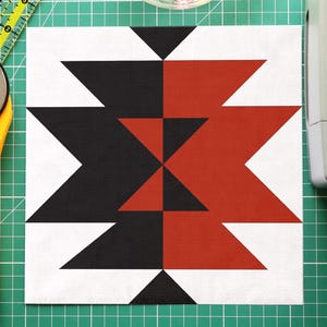 May include: A square quilt block with a geometric design in black, red, and white. A yellow rotary cutter, a clear bowl of colorful pins, and a green cutting mat are also visible. A sewing machine is in the background.