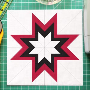Puede incluir: Un bloque de patchwork con un diseño de estrella en rojo, negro y blanco, junto con un cortador rotatorio amarillo, una esterilla de corte verde, un cuenco con alfileres de colores y una máquina de coser.