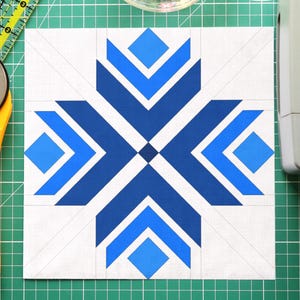 May include: A white and blue quilt block with a geometric design, a yellow rotary cutter, a clear bowl of colorful pins, a green cutting mat, and a clear ruler. Sewing and quilting supplies.