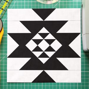 Pode incluir: Um bloco de colcha geométrico em preto e branco com um cortador rotativo, uma régua, uma máquina de costura e alfinetes em uma esteira de corte verde. O bloco de colcha apresenta um padrão de triângulo repetitivo.