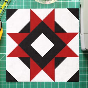 May include: A quilt block with a geometric star design in red, black, and white, surrounded by sewing tools. A yellow rotary cutter, a clear bowl of colorful pins, and a green cutting mat with a ruler are also visible.