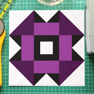 Könnte beinhalten: Ein Patchwork-Quiltblock mit einem geometrischen Design in Lila, Schwarz und Weiß. Ein gelber Rollschneider, eine transparente Schüssel mit bunten Stecknadeln und ein grünes Lineal liegen auf einer grünen Schneidematte. Eine Nähmaschine ist im Hintergrund.