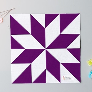 Puede incluir: Un bloque de edredón cuadrado con un diseño de estrella en blanco y morado. Accesorios de costura, incluyendo alfileres con cabezas de botón pastel y pequeños clips, rodean el bloque. El logotipo de The Cozy Quilt Studio es visible.