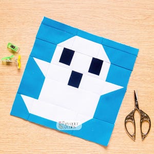 May include: A square quilt block featuring a white ghost shape with two black square eyes and a black square mouth, set against a bright blue background. The design includes a small logo reading The Cozy Quilt Studios at the bottom centre.