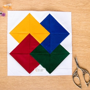 Puede incluir: Un bloque de edredón cuadrado con fondo blanco y cuatro grandes rombos en amarillo, azul, rojo y verde oscuro. Una pequeña etiqueta blanca muestra el texto The Cozy Quilt Studios. Tres pequeñas pinzas de costura de plástico y tijeras de metal.