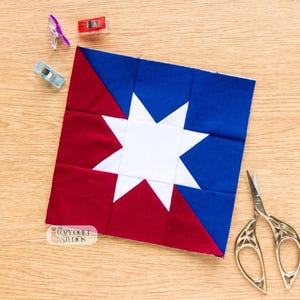 May include: A square fabric quilt block featuring a central white eight-pointed star set against a background of deep red and royal blue geometric sections. The block includes a small logo reading The Cozy Quilt Studios, three plastic sewing clips, and silver scissors.
