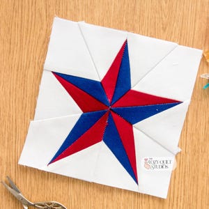 May include: A white, red, and blue star quilt block with a star design. The star is made of red and blue fabric, and the background is white. The block is on a wooden surface, with sewing clips and scissors nearby. The Cozy Quilt Studios logo is visible.