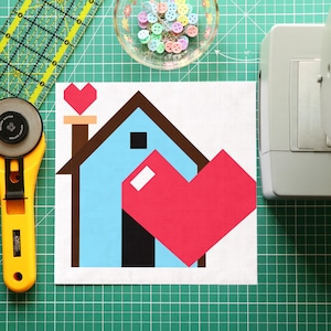 May include: A fabric block with a house and heart design, featuring blue, brown, black, and red shapes. A yellow rotary cutter, a green ruler, and a bowl of colorful pins are also visible on a green cutting mat.