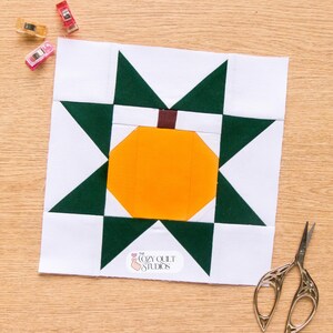 Puede incluir: Un bloque de edredón cuadrado con una forma de calabaza naranja central con un tallo marrón, rodeado por un patrón de estrella verde oscuro sobre un fondo blanco. El bloque incluye el texto The Cozy Quilt Studios. Se ven tres clips de costura de plástico y tijeras de metal.