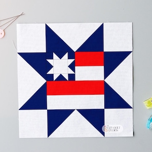 Pode incluir: Um bloco de colcha quadrado com um design de estrela em branco, azul marinho e vermelho. O bloco apresenta uma estrela central e listras, com o texto "The Cozy Quilt Studio" no canto inferior direito. Alfinetes e clipes de costura no fundo.
