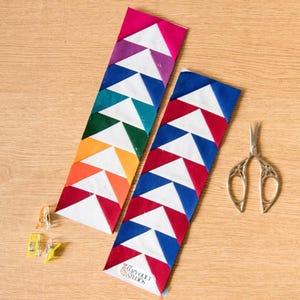 May include: Two colorful fabric bookmarks with a geometric triangle pattern in red, orange, yellow, green, blue, and purple. A pair of decorative scissors and sewing clips are also on the wooden surface. The text "Cozy Quilt Studios" is visible.