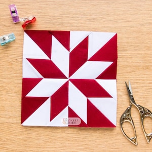 May include: A square fabric quilt block featuring a red and white eight-pointed star pattern. The block includes a small logo reading The Cozy Quilt Studios. Nearby are three plastic sewing clips in red and blue, and a pair of silver metal scissors.