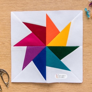 Puede incluir: Un bloque de edredón cuadrado con un diseño de molinete vibrante hecho de triángulos de tela multicolores en rojo, naranja, amarillo, verde oscuro, verde azulado, azul, morado y magenta, sobre un fondo blanco con una pequeña etiqueta de logotipo.