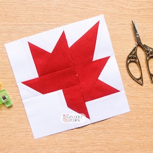 May include: A square fabric quilt block featuring a red maple leaf design on a white background. The block includes the text The Cozy Quilt Studios. Beside the block are silver metal embroidery scissors and two small green and yellow plastic clips.