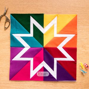 May include: A colorful star quilt block with teal, green, yellow, orange, red, pink, and purple sections. White fabric forms the star design. A pair of silver scissors and several small sewing clips are on a wooden surface.