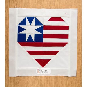May include: A square fabric quilt block featuring a heart shape composed of red and white horizontal stripes on the right and a blue square with a white eight-pointed star on the left. The block has a white border and a small logo reading The Cozy Quilt Studios.