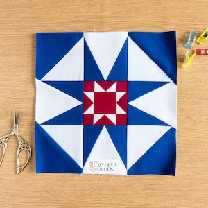 Peut inclure: Un bloc de courtepointe carré avec un motif d'étoile rouge, blanc et bleu. Le bloc présente un carré rouge central entouré de triangles blancs et bleus. Des ciseaux et des pinces à quilting sont sur une surface en bois.
