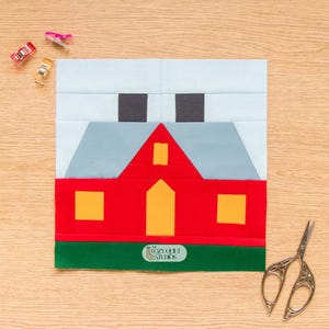 May include: A fabric quilt block featuring a red house with a gray roof, yellow windows, and a yellow door against a light blue sky. Two black squares represent chimneys. A green strip forms the ground, labeled with The Cozy Quilt Studios logo.