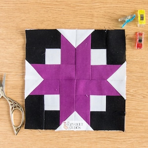 May include: A square quilt block with a purple star design, black and white accents, and a small logo in the corner. The block is surrounded by sewing clips in yellow, red, and blue, and a pair of silver scissors. The items are on a wooden surface.