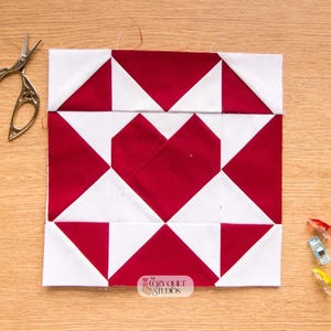 Op de afbeelding: Een vierkant quiltblok met een rood hartontwerp in het midden, omringd door rode en witte driehoeken. Het blok ligt op een houten oppervlak, met een schaar en quiltclips in de buurt. Het Cozy Quilt Studios logo is zichtbaar.