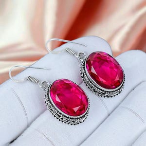 Puede incluir: Un par de pendientes plateados con grandes piedras preciosas rosas intensas de corte ovalado. Las piedras están engastadas en biseles ornamentados y detallados. Los pendientes tienen cierre de gancho.