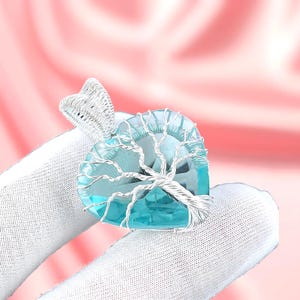 Może przedstawiać: Zawieszka w kształcie serca z jasnoniebieskim kamieniem szlachetnym, misternie owinięta srebrnym drutem, przypominającym drzewo. Zawieszka jest zawieszona na srebrnym uchwycie. Tło jest w delikatnym różu.