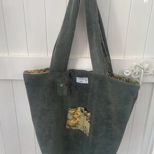 Peut inclure: Sac fourre-tout en velours côtelé vert olive avec de longues anses. Il comporte une petite poche plaquée avec un motif camouflage et une étiquette avec du texte. Le sac a une doublure à motifs.
