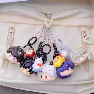 May include: A collection of colorful character keychains with black keyrings and silver chain attachments. The keychains feature various anime-style characters with detailed facial features and hair, attached to a white bag.