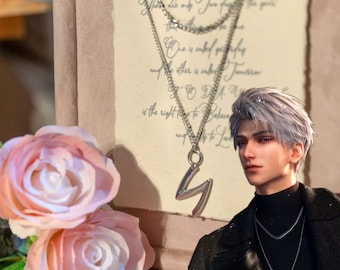 May include: A silver-toned necklace with a stylized "S" pendant hangs in front of a person. The person is wearing a black turtleneck and jacket. A second necklace with a "Z" pendant is also visible. Soft pink roses and a handwritten note are in the background.
