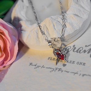 May include: A silver necklace with a pendant featuring a red gemstone. The pendant is encased in a detailed silver design. The necklace is displayed on a white surface with a pink rose in the background. Text on the surface reads "PLEASE TAKE ONE".
