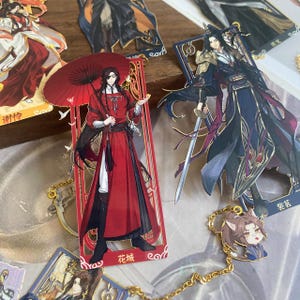 Pode incluir: Uma coleção de marcadores e berloques de personagens em estilo anime. O marcador central apresenta um personagem com uma longa túnica vermelha, segurando um guarda-chuva vermelho. Outros marcadores e berloques retratam personagens com trajes detalhados, com detalhes dourados e desenhos intrincados.