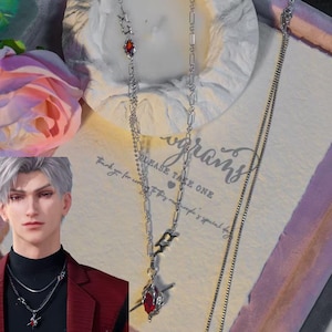 May include: A silver necklace with a red gemstone pendant. The necklace has a delicate chain design with a stylized charm and a teardrop-shaped red stone. The image also includes a second, simpler chain.