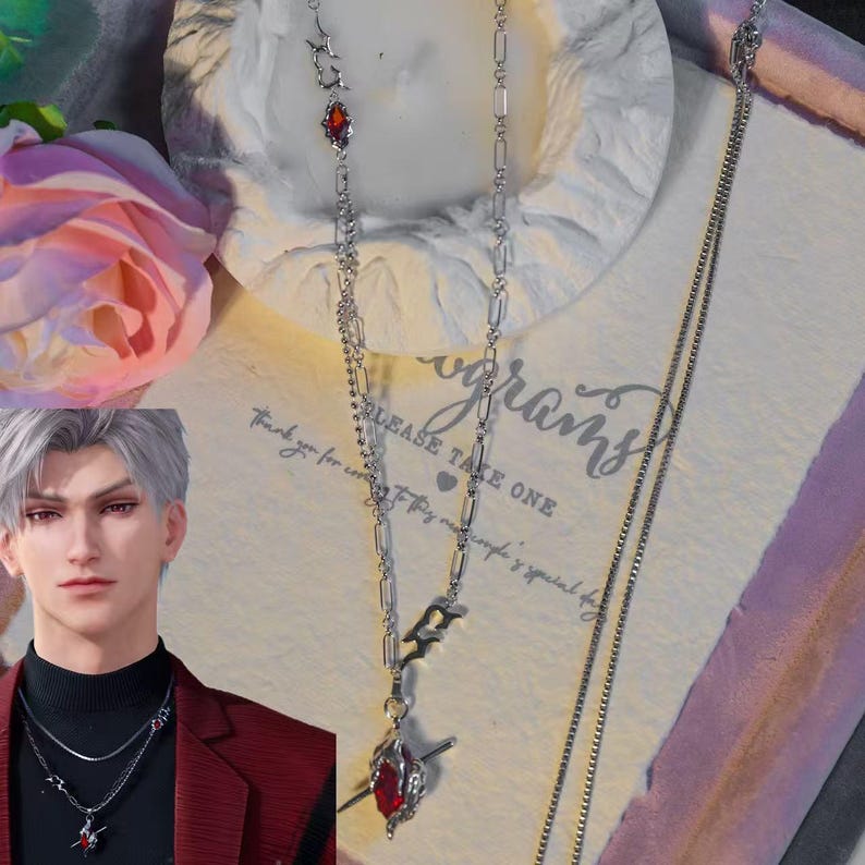 May include: A silver necklace with a red gemstone pendant. The necklace has a delicate chain design with a stylized charm and a teardrop-shaped red stone. The image also includes a second, simpler chain.