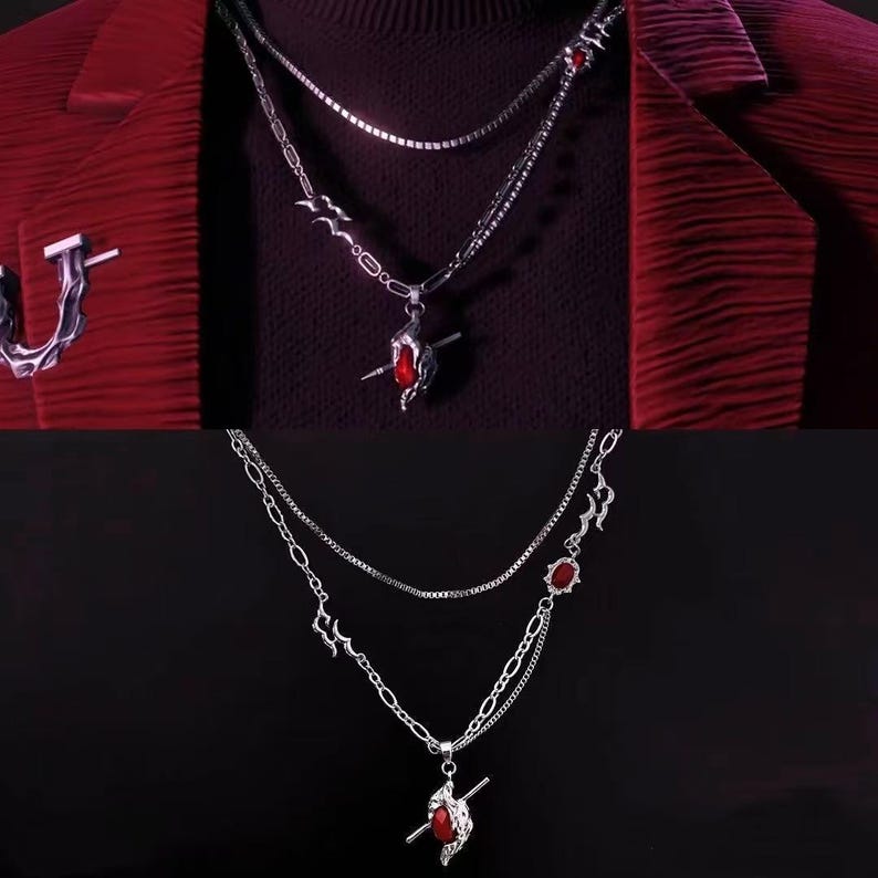 May include: Silver-toned layered necklace with a pendant featuring a red gemstone and a small silver bar. The necklace has a delicate chain design with additional red gemstones. The image shows the necklace against a dark background.