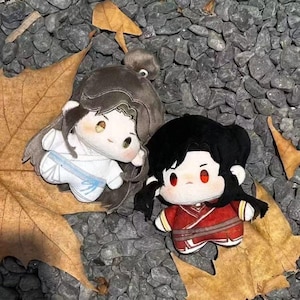 May include: Two plush dolls, one in white with a blue sash and the other in red, are displayed on a bed of gray stones and fallen leaves. The dolls have large eyes and black hair, with one doll's hair tied in a bun.