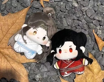 Può includere: Due bambole di peluche, una bianca con una fascia blu e l'altra rossa, sono esposte su un letto di pietre grigie e foglie cadute. Le bambole hanno grandi occhi e capelli neri, con i capelli di una bambola legati in uno chignon.