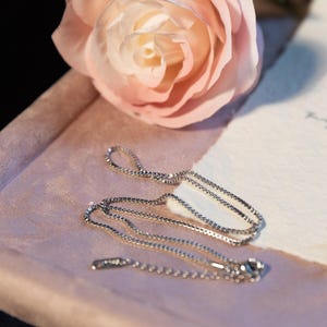 May include: A delicate silver necklace with a fine chain and a small pendant. The necklace is displayed on a pink surface with a soft pink rose in the background. The overall aesthetic is elegant and feminine.