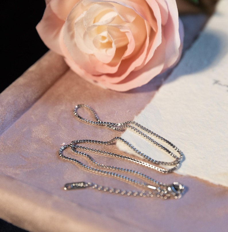 May include: A delicate silver necklace with a fine chain and a small pendant. The necklace is displayed on a pink surface with a soft pink rose in the background. The overall aesthetic is elegant and feminine.