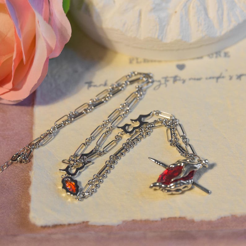 May include: A silver necklace with a flame-like design and a red gemstone pendant. The necklace is on a cream-colored surface with a pink rose in the background. The pendant has a unique, artistic design.
