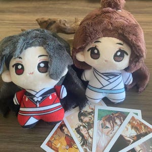 May include: Two plush dolls with large eyes and soft hair, one in a red and white outfit, the other in white with blue accents. Several small cards with illustrations of anime characters are in front of the dolls.