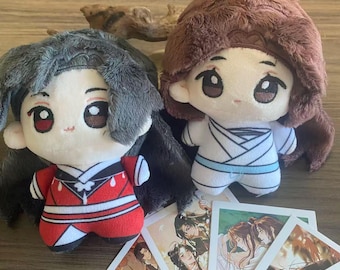Può includere: Due bambole di peluche con grandi occhi e capelli morbidi, una in un completo rosso e bianco, l'altra in bianco con accenti blu. Diverse piccole carte con illustrazioni di personaggi anime sono di fronte alle bambole.